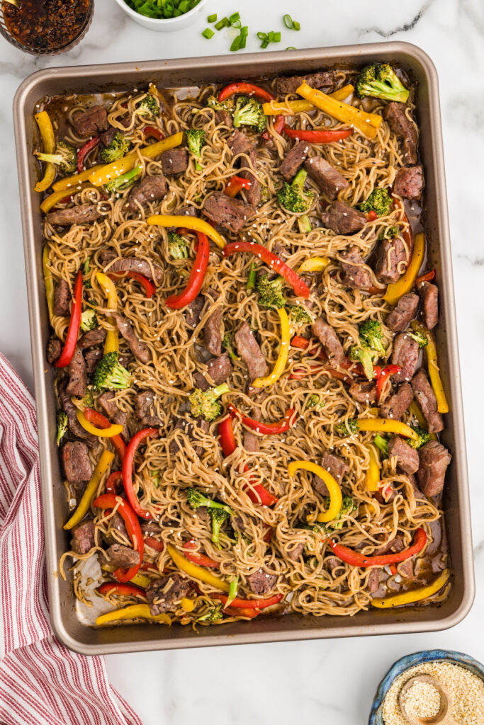 Sheet pan of beef and ramen noodles with broccoli and bell peppers, garnished with sesame seeds.