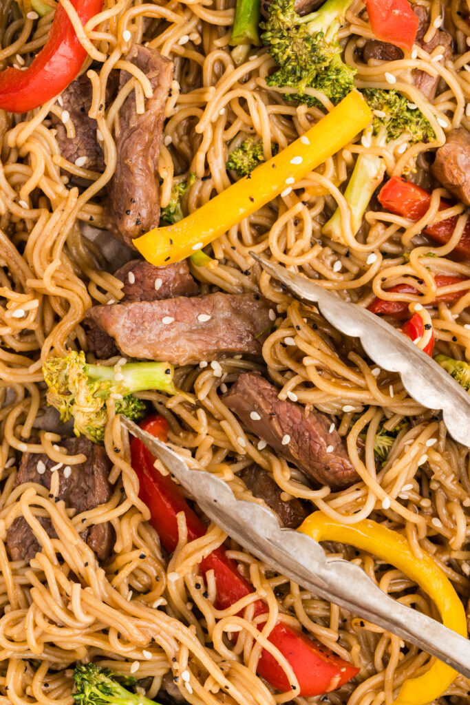 Close-up of beef and ramen noodles with broccoli and bell peppers, sprinkled with sesame seeds.