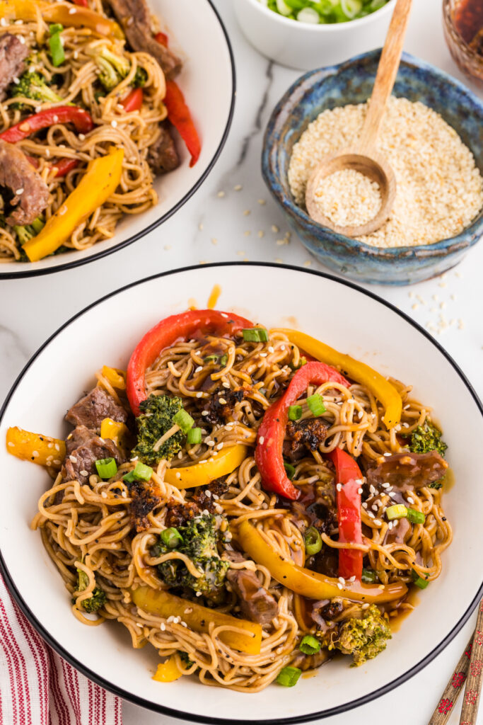 Beef and ramen noodles with broccoli and bell peppers in a bowl, topped with sesame seeds and green onions.