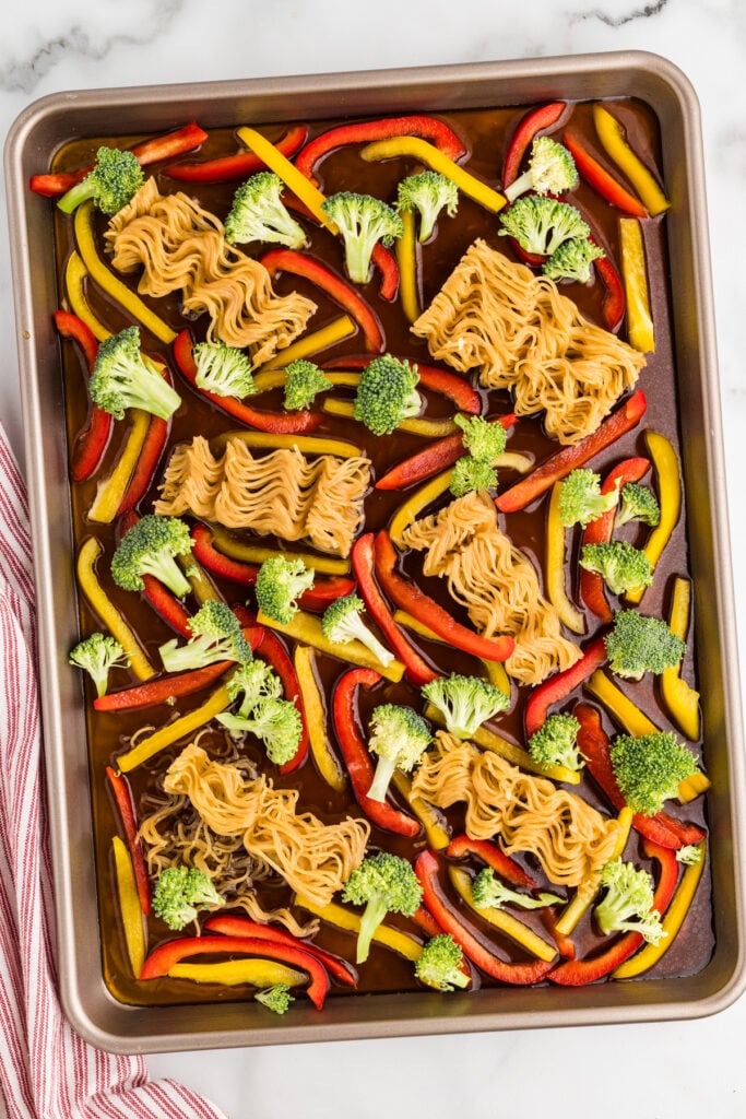 Sheet pan with ramen noodles, broccoli, and bell peppers in sauce.