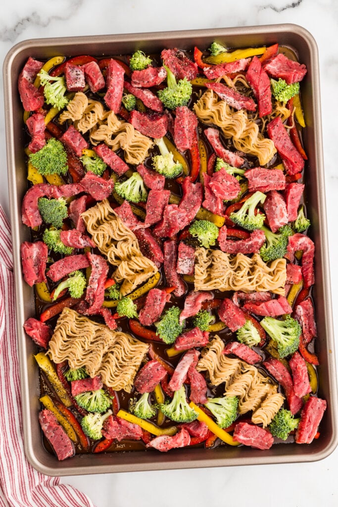 Sheet pan with raw beef, dry ramen noodles, broccoli, and bell peppers in sauce.