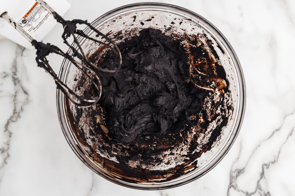 Dark brownie batter in a glass mixing bowl with electric mixer beaters on a marble countertop.