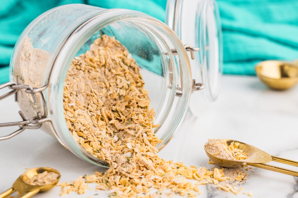 Homemade onion soup mix spills from a glass jar beside gold measuring spoons.