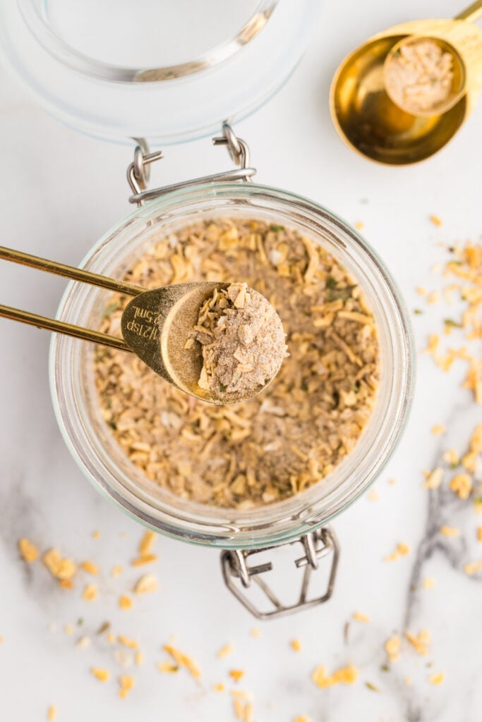 Gold measuring spoons scoop homemade onion soup mix from a glass jar on a marble surface.