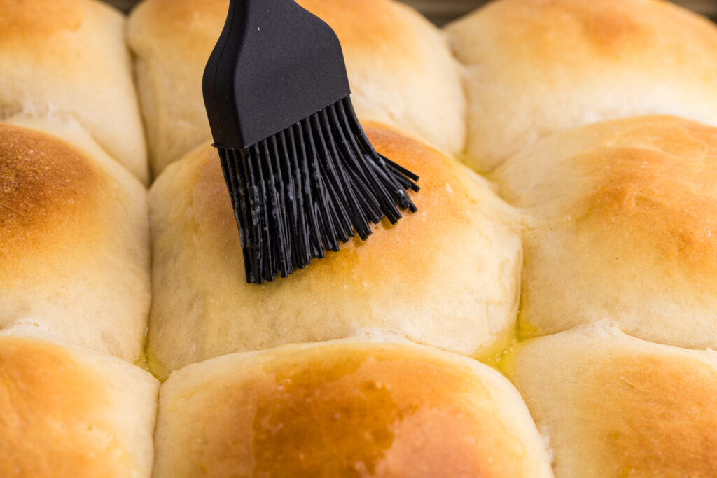 Pastry brush spreading butter over warm potato dinner rolls.