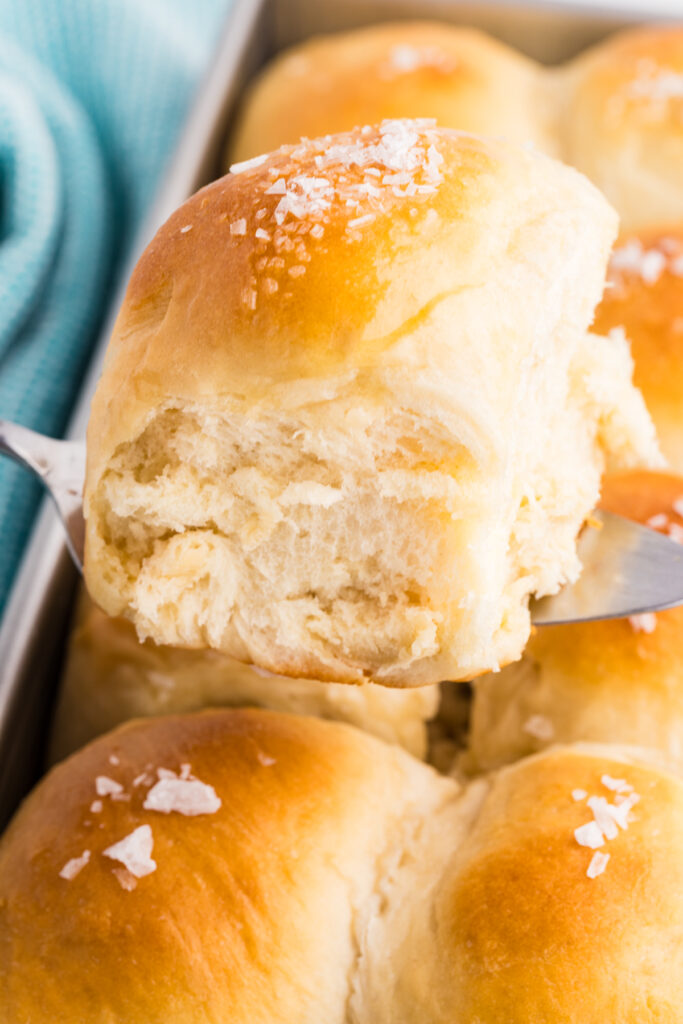 Close-up of a potato dinner roll lifted from a pan with flaky salt on top.