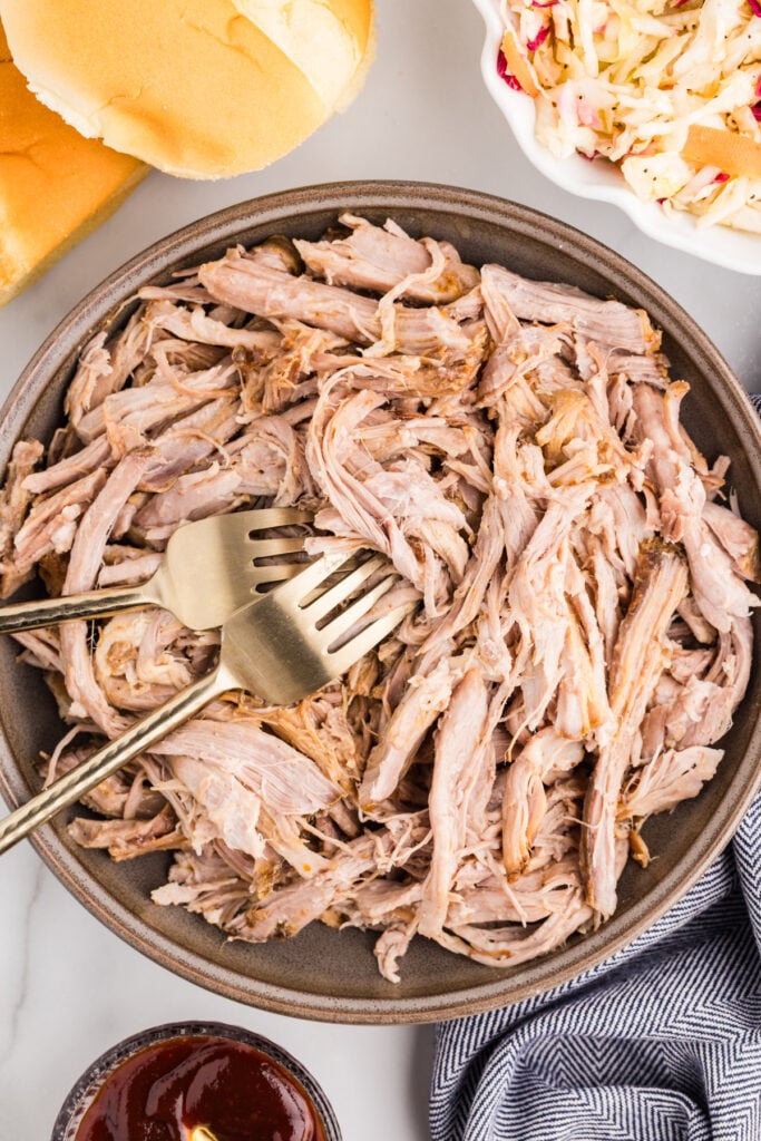 Bowl of shredded pulled pork with buns, coleslaw, barbecue sauce, and two forks.