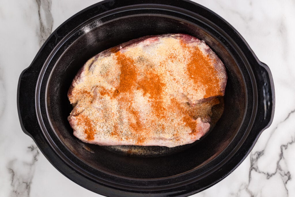 Raw pork shoulder coated with seasoning sits inside a black slow cooker insert on a white marble countertop.