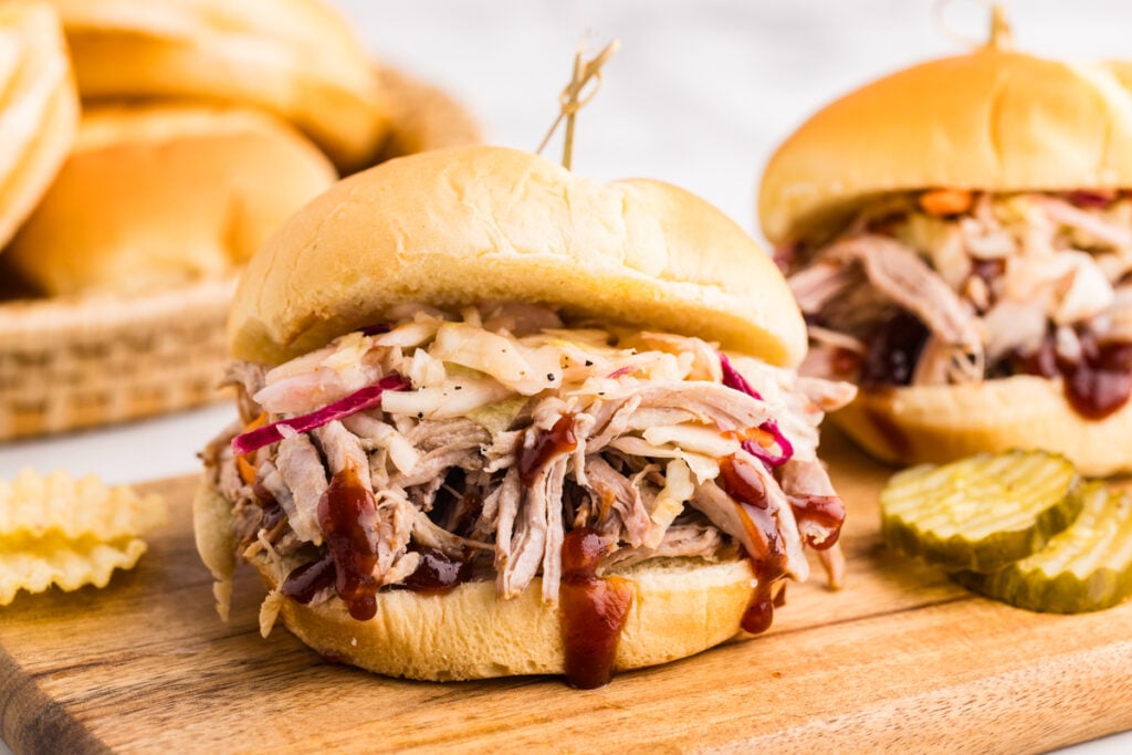 A close-up of a pulled pork sandwich on a soft hamburger bun, topped with creamy coleslaw and drizzled with barbecue sauce.
