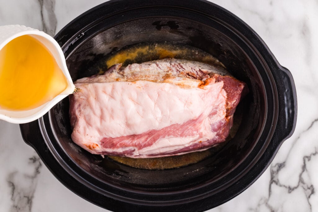 Chicken broth is poured over a seasoned pork shoulder in a black slow cooker.