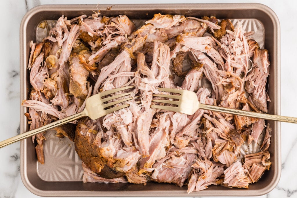Shredded slow-cooked pork fills a sheet pan while two forks pull apart the tender meat.