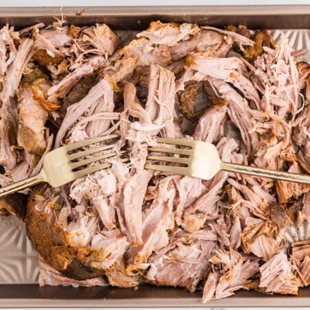 Shredded slow-cooked pork is spread across a sheet pan with two forks pulling apart the meat.