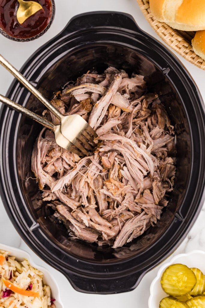 Shredded pulled pork sits in a black slow cooker with buns, barbecue sauce, coleslaw, and pickles nearby.