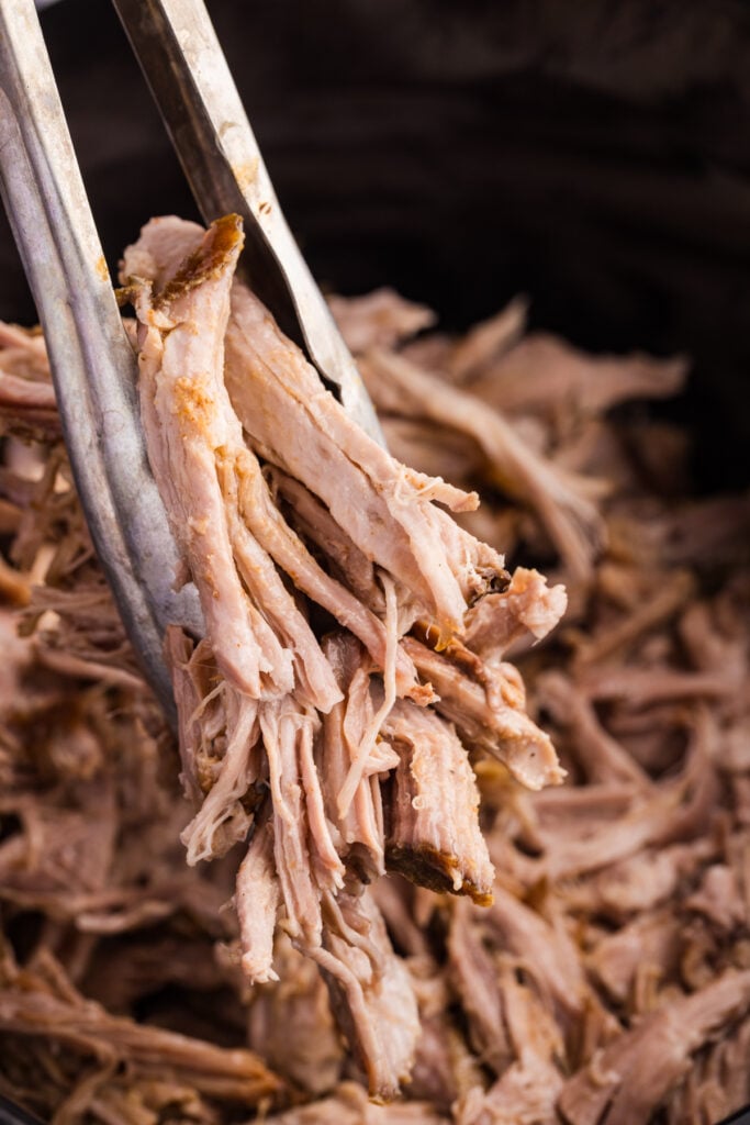 A close-up of metal tongs lifting a bundle of tender shredded pulled pork from a slow cooker.