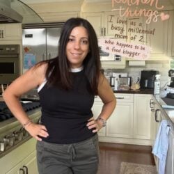 A smiling woman with dark hair stands with hands on her hips in a kitchen with cream cabinets and stainless steel appliances. Handwritten text overlaid on the top right reads: "kitchen things I love as a busy millennial mom of 2... who happens to be a food blogger.