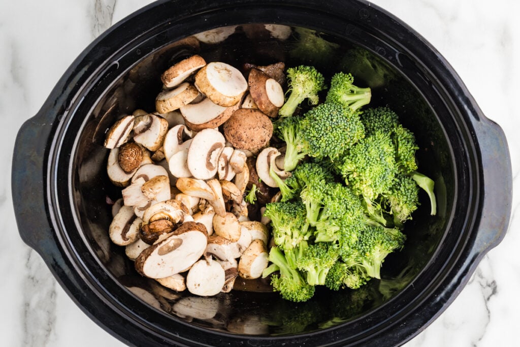Sliced mushrooms and broccoli florets added to a black slow cooker.