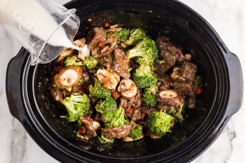 Cornstarch mixture being poured into a slow cooker filled with beef, broccoli, and sliced mushrooms.