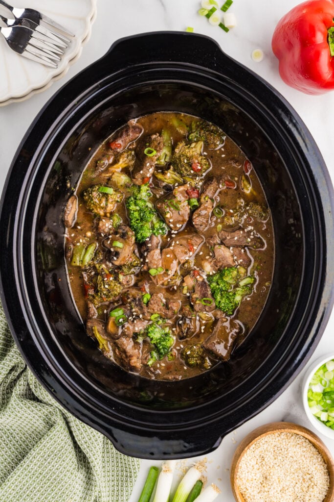 Overhead view of slow cooker pepper steak in a black crockpot surrounded by green onions, sesame seeds, and a red bell pepper.