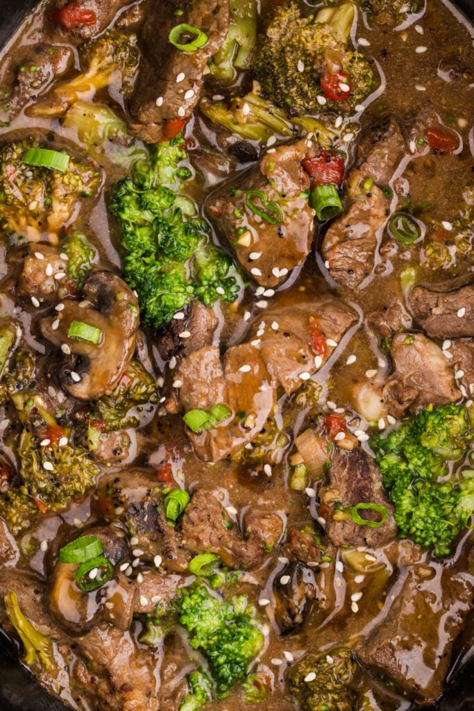 Slow cooker pepper steak with broccoli, mushrooms, and red peppers in a savory brown sauce, topped with sesame seeds and green onions.