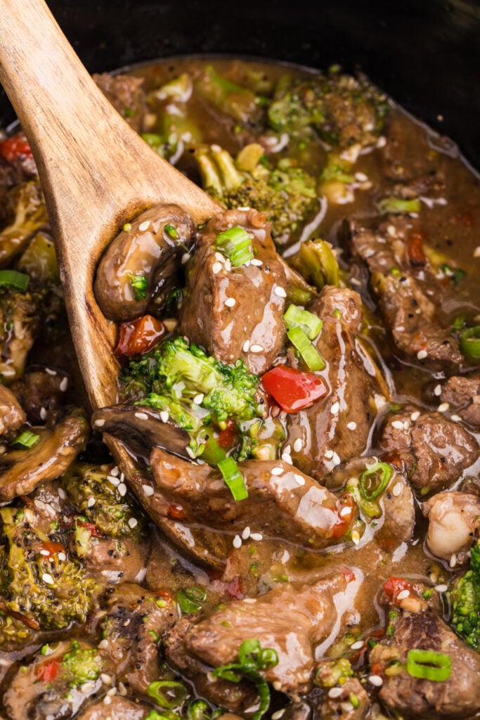 Wooden spoon lifting slow cooker pepper steak with broccoli, mushrooms, and red peppers from a crockpot filled with savory brown sauce.
