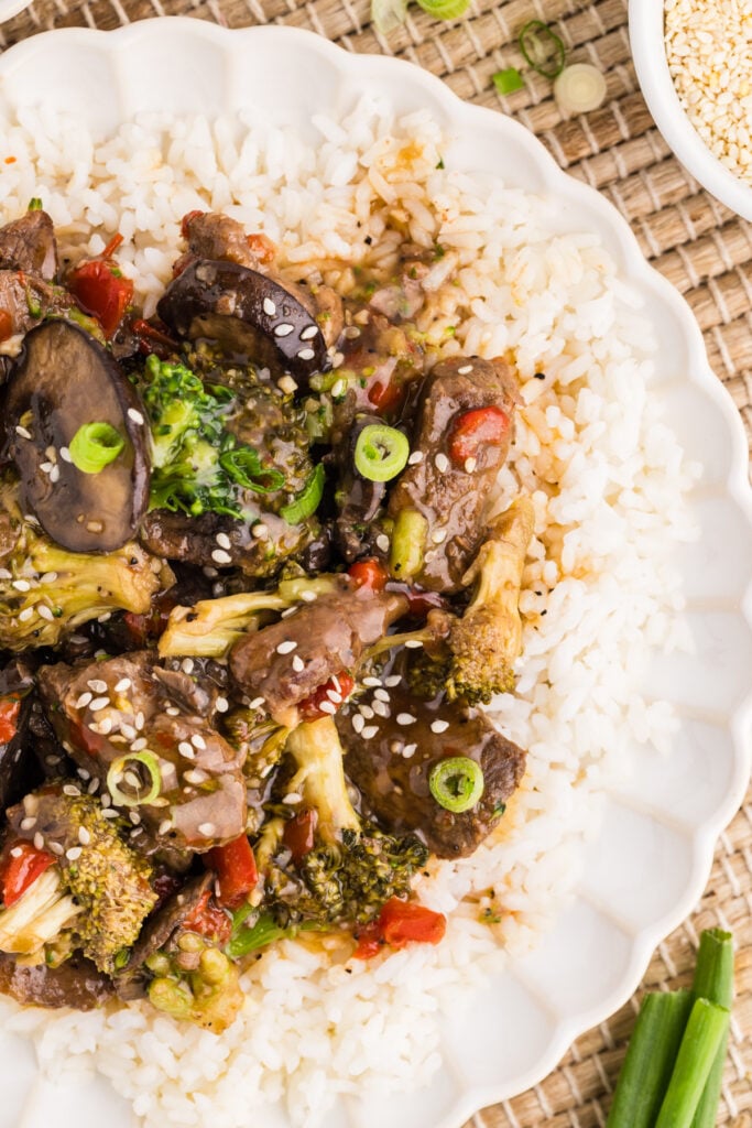 Slow cooker pepper steak with broccoli, mushrooms, and red peppers served over white rice and topped with sesame seeds and green onions.