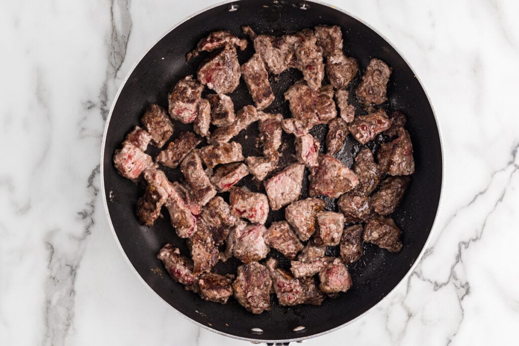 Chunks of browned beef cooking in a skillet on a white marble surface.