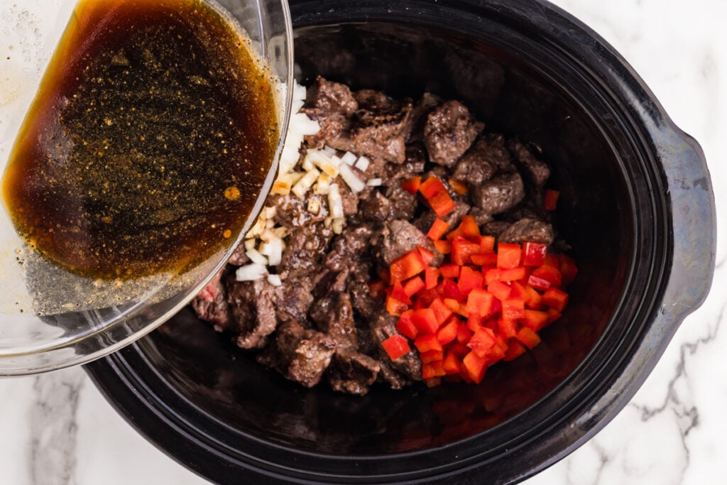Sauce being poured over browned beef, diced red peppers, and onions in a slow cooker.
