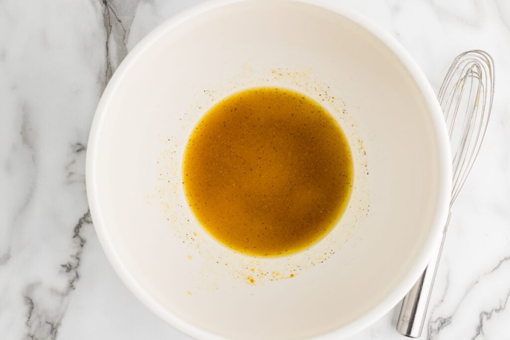 Vinegar dressing with black pepper in a white mixing bowl beside a metal whisk.