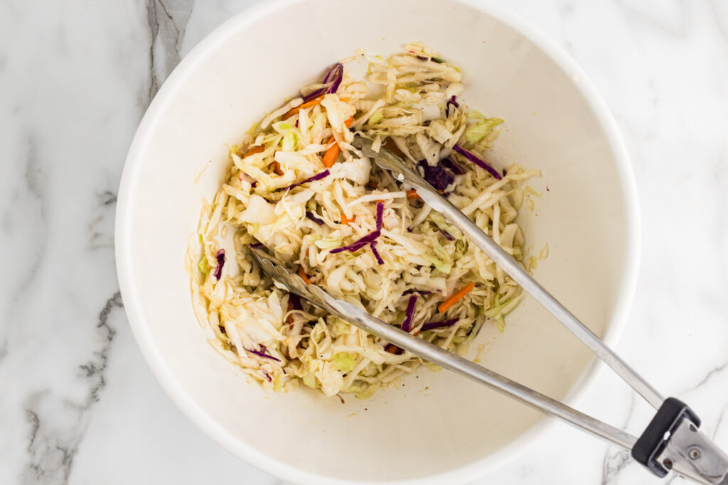 Coleslaw mix tossed in a white bowl with metal tongs.