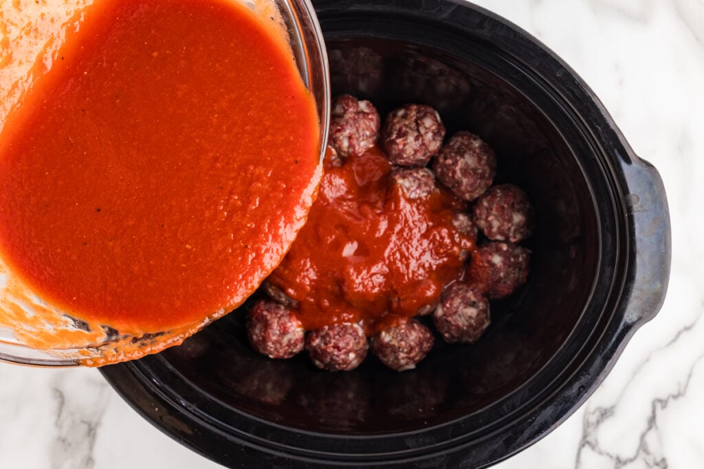 Tomato sauce being poured over raw porcupine meatballs in a slow cooker.