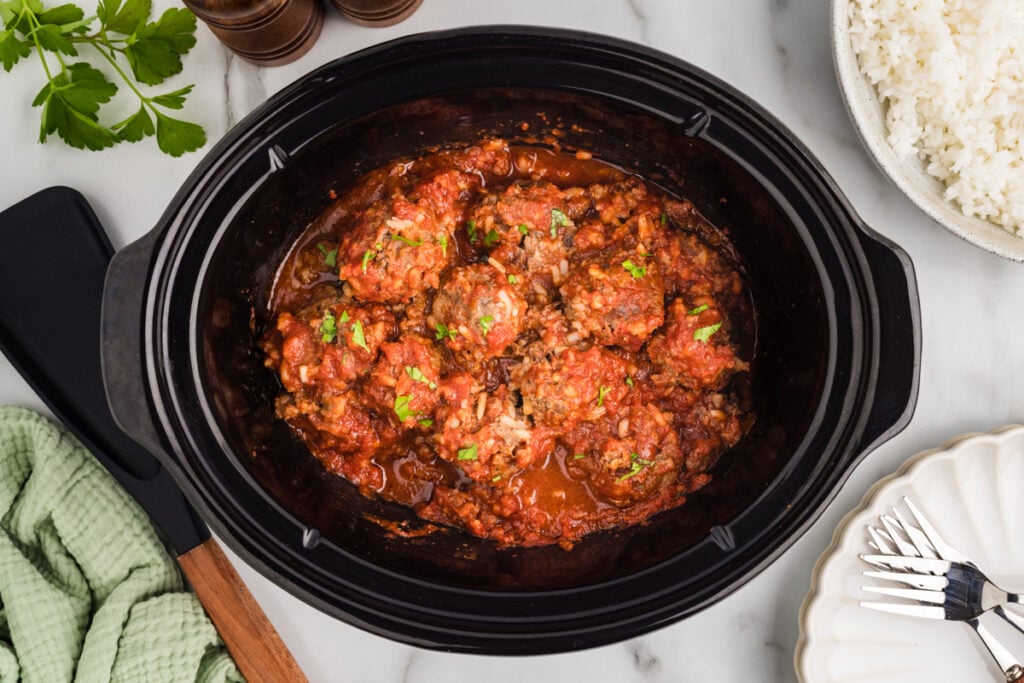 Overhead view of cooked porcupine meatballs in a black slow cooker with serving dishes nearby.