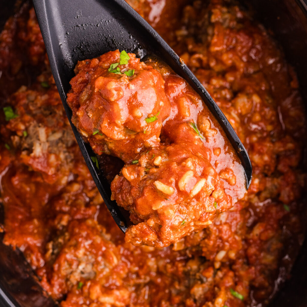 Black serving spoon lifting two porcupine meatballs coated in thick tomato sauce from a slow cooker.