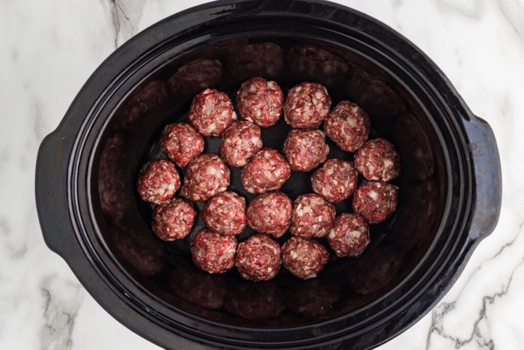 Raw porcupine meatballs arranged in a single layer inside a black slow cooker.